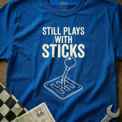 Blue t-shirt with "Still Plays With Sticks" text and a manual transmission shifter graphic.