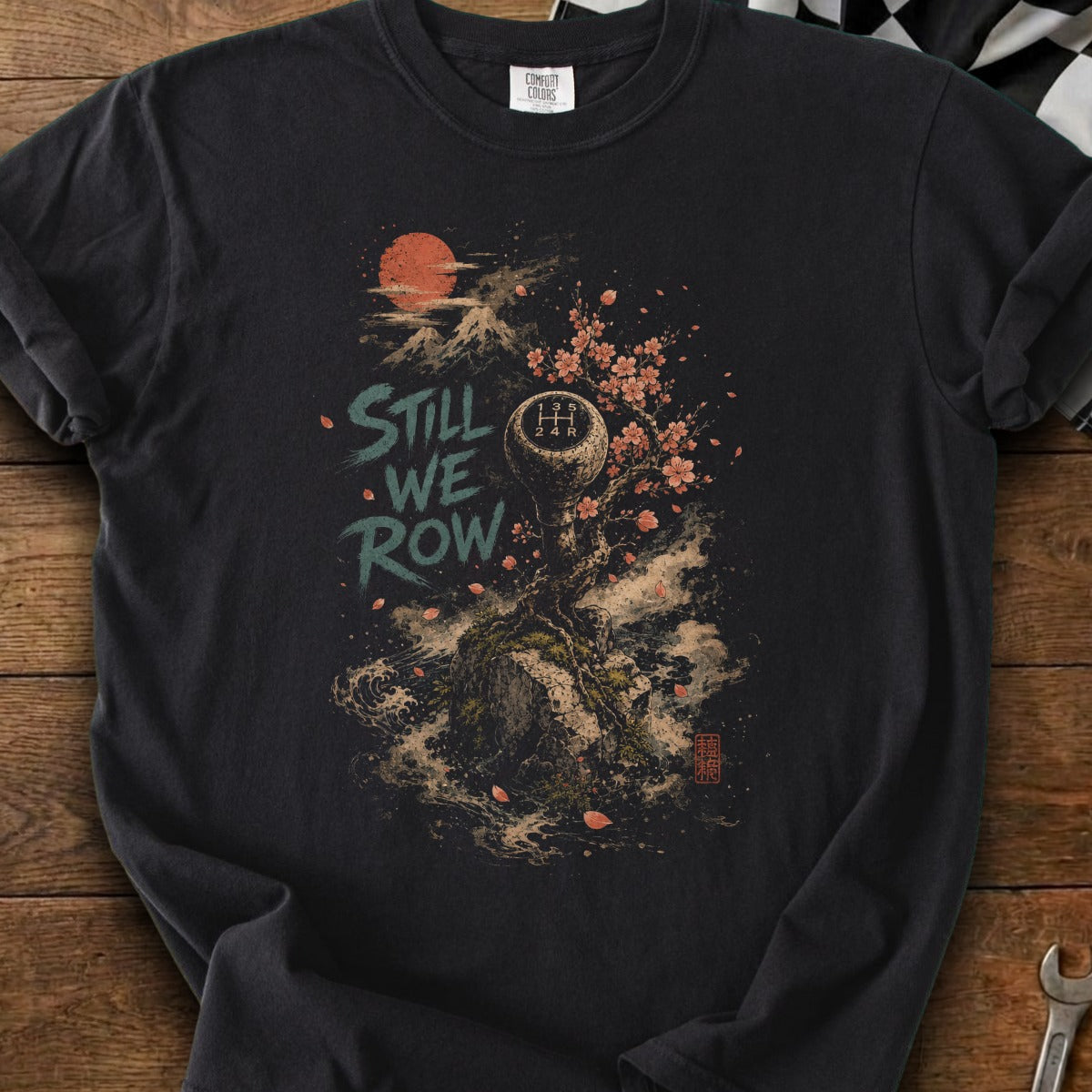 Graphic t-shirt with "Still We Row" text, gear shift, cherry blossoms, and a mountain sun.