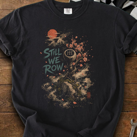 Graphic t-shirt with "Still We Row" text, gear shift, cherry blossoms, and a mountain sun.