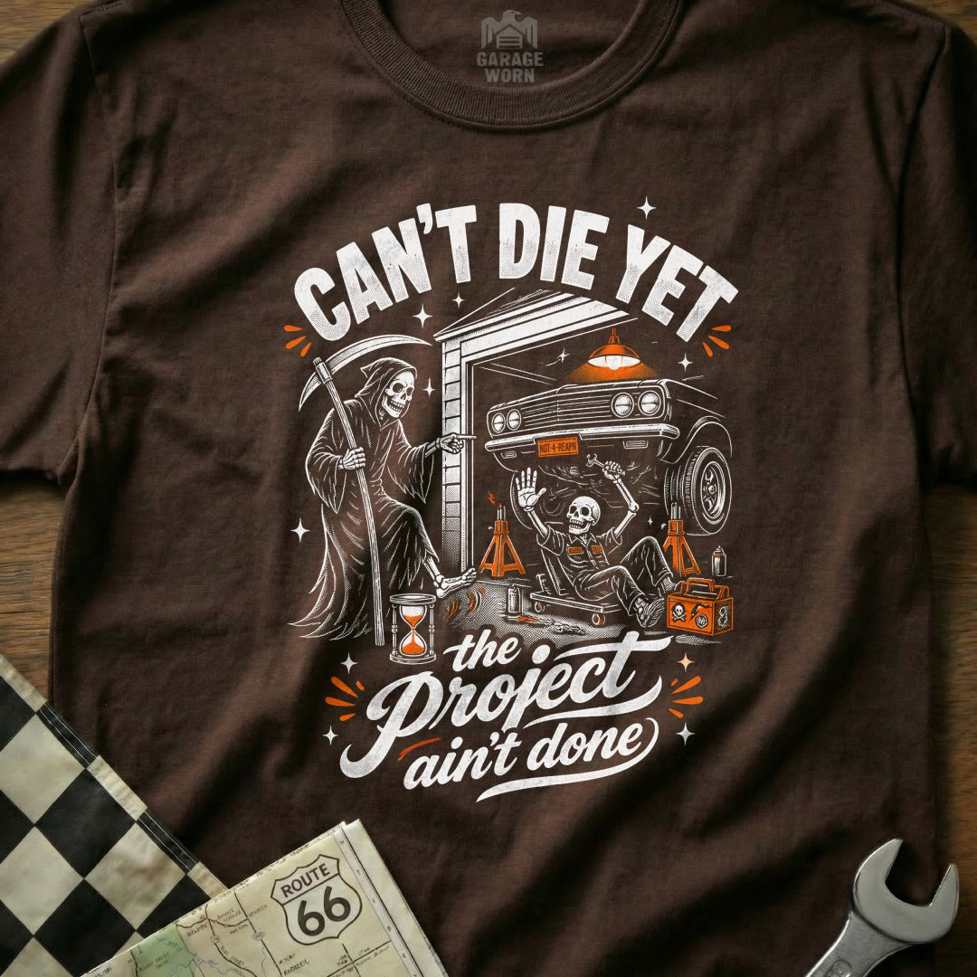 Grim Reaper points to skeleton working on car, "Can't Die Yet, The Project Ain't Done".
