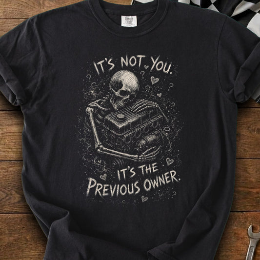 Black t-shirt with skeleton holding car engine design and text "It's not you. It's the previous owner."