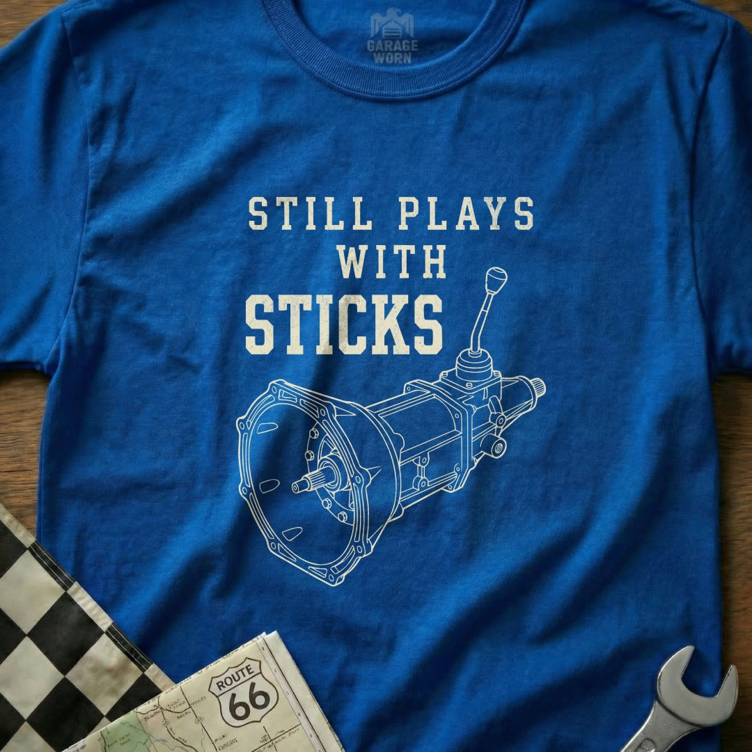Blue t-shirt with white text "STILL PLAYS WITH STICKS" and gear shifter diagram.