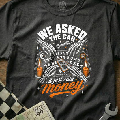 Skeleton hands use a Ouija board on a t-shirt with "We Asked The Car It Just Said Money."