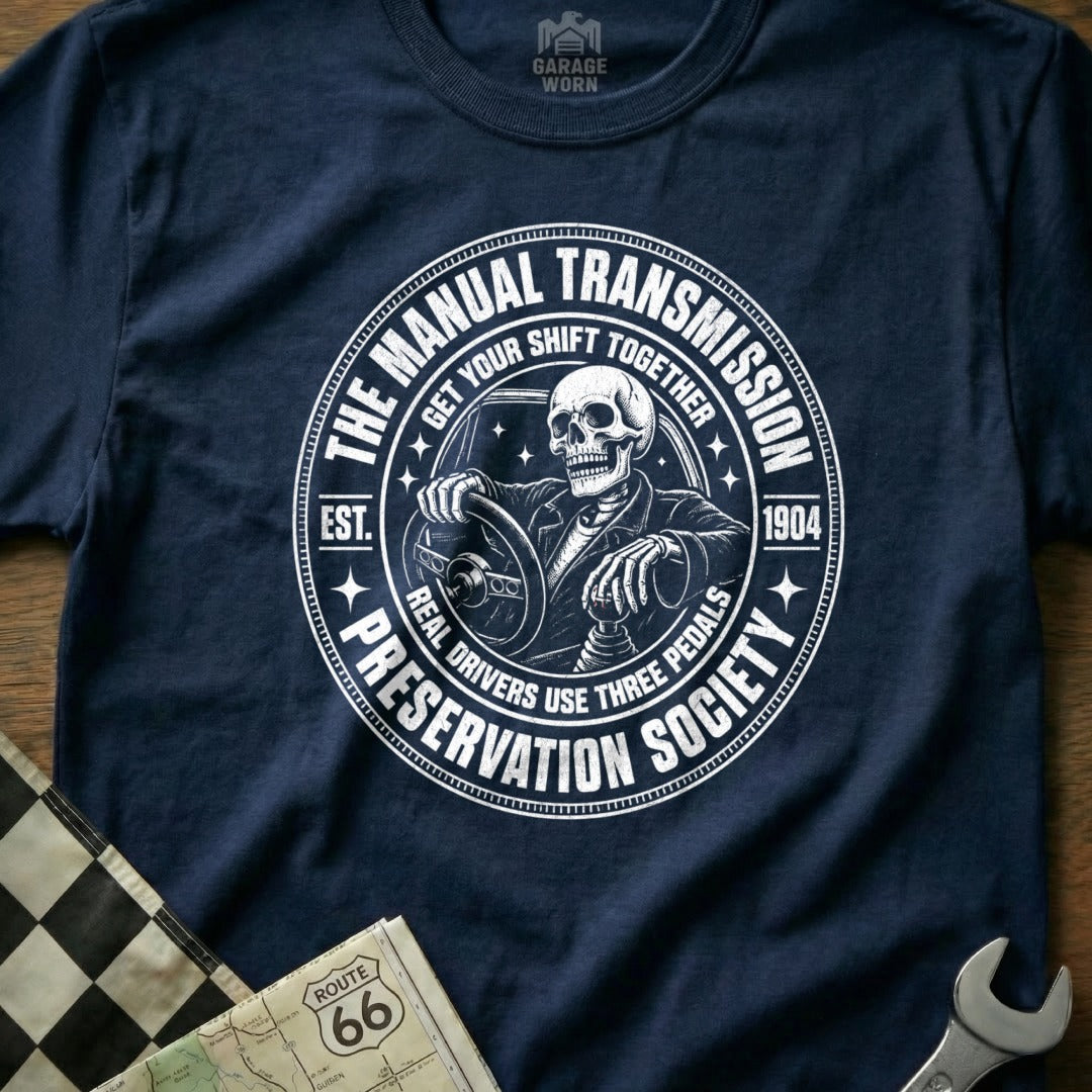 Navy t-shirt featuring a skeleton driving a manual transmission with text "The Manual Transmission Preservation Society".
