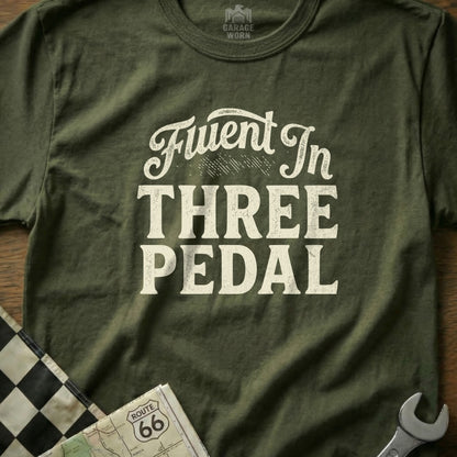 Green t-shirt with "Fluent In Three Pedal" text and checkered flag detail.