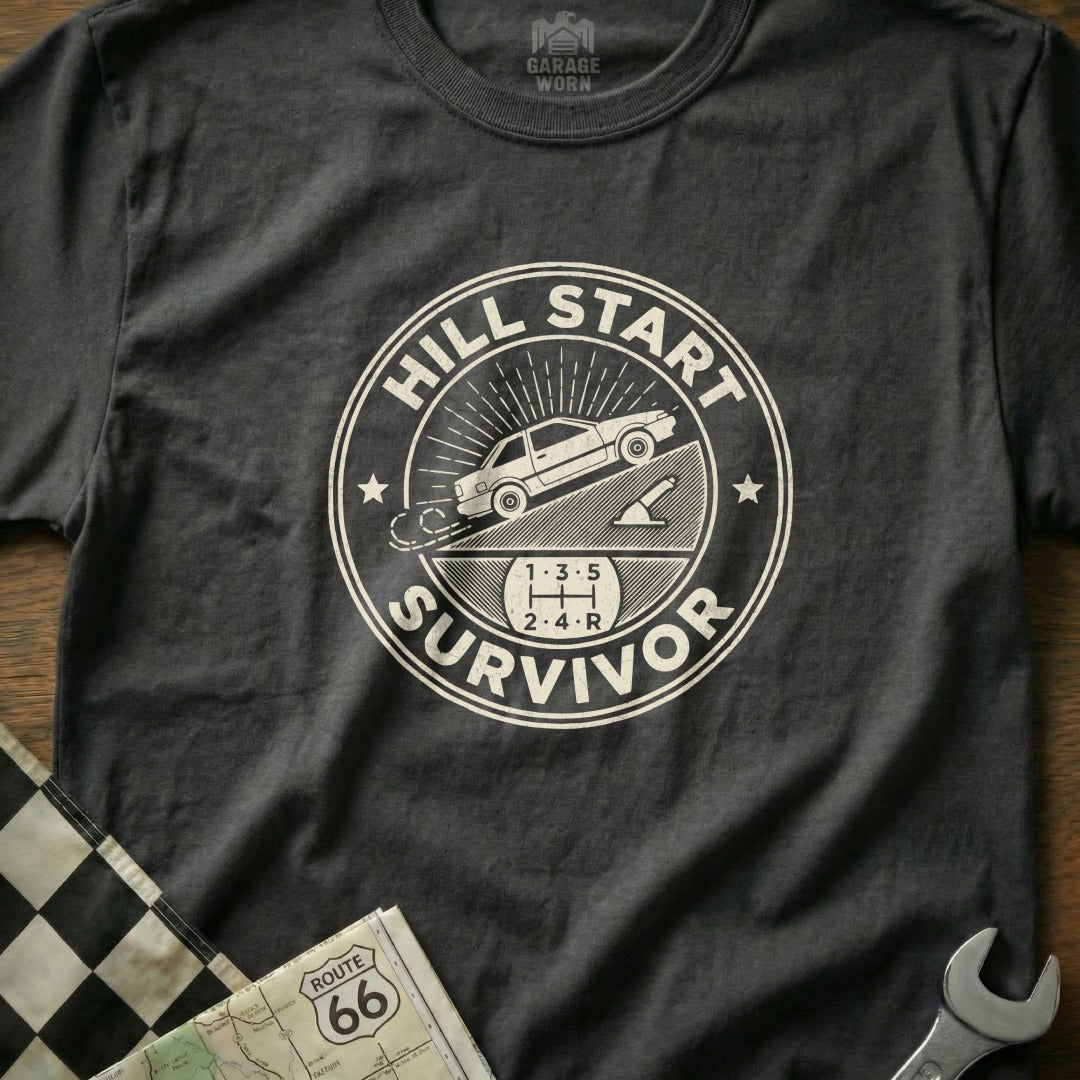 Dark t-shirt with "Hill Start Survivor" graphic featuring a car on an incline and gear shift pattern.