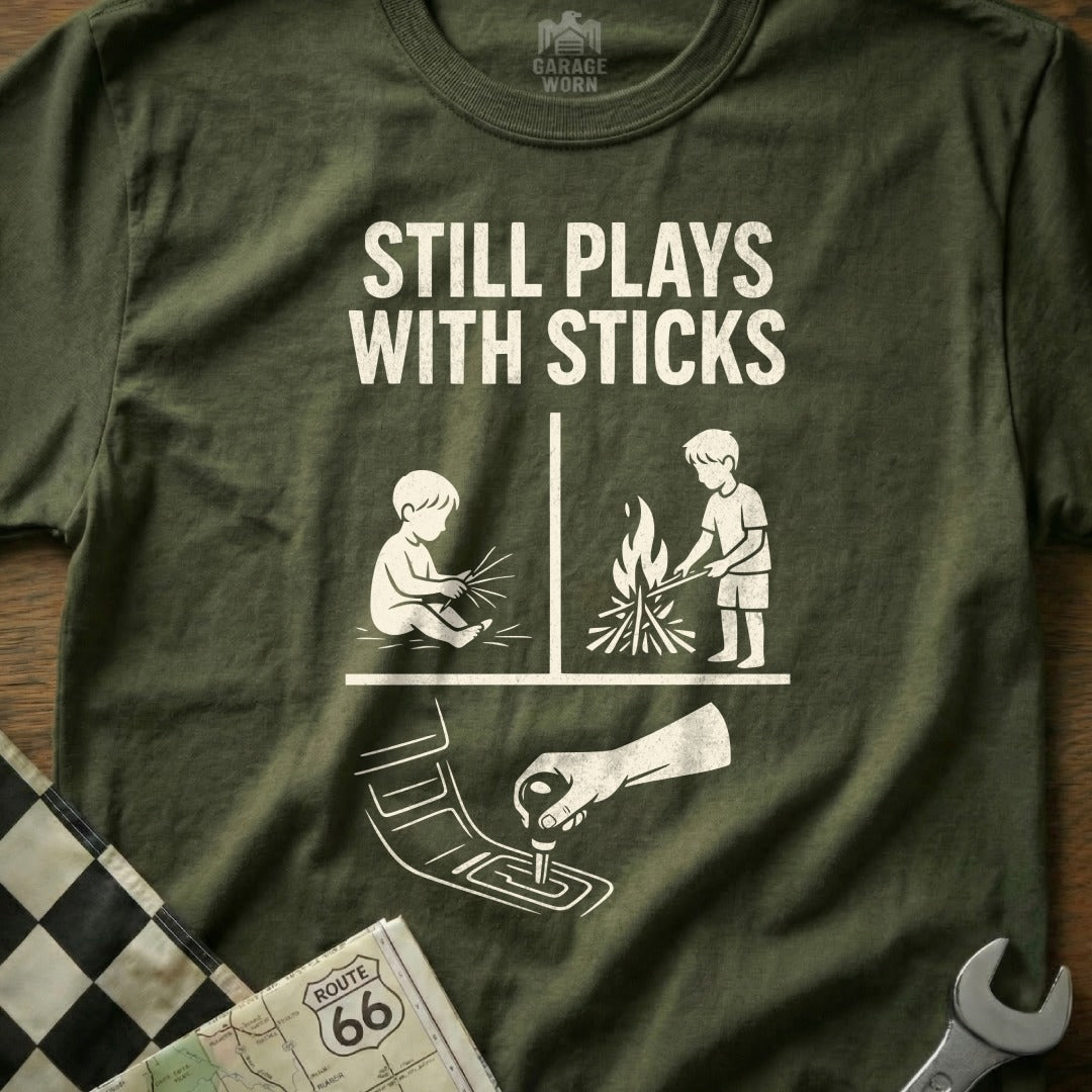 Green t-shirt with "Still Plays With Sticks" text and graphics of a child playing with sticks and a campfire.
