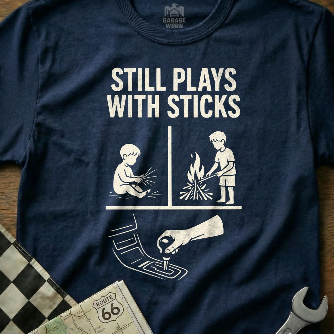 Navy t-shirt with "STILL PLAYS WITH STICKS" and illustrations of child and campfire.