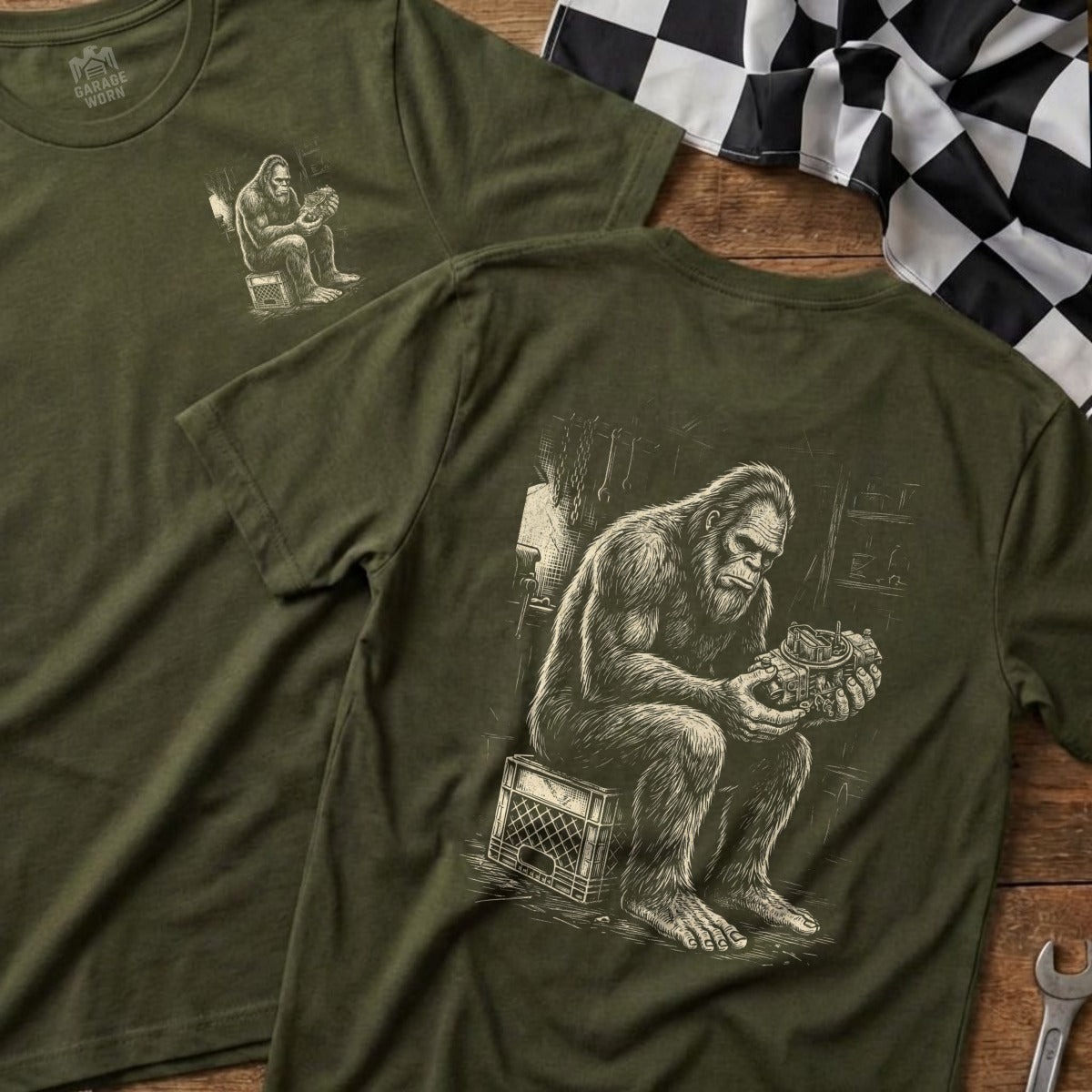 Bigfoot on a crate examines a carburetor, printed on an olive green t-shirt.