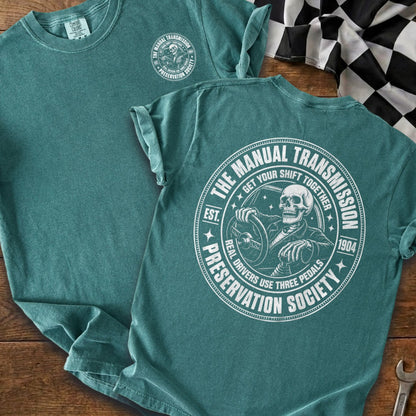 Green t-shirt with white graphic of skeleton driving, with text "The Manual Transmission Preservation Society".