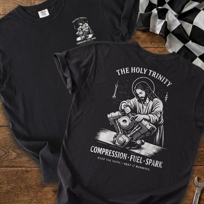 Black t-shirt with Jesus working on a motorcycle engine, text "The Holy Trinity Compression Fuel Spark".