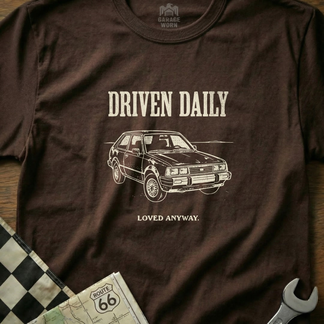 Brown t-shirt with "DRIVEN DAILY LOVED ANYWAY" text and a car graphic.