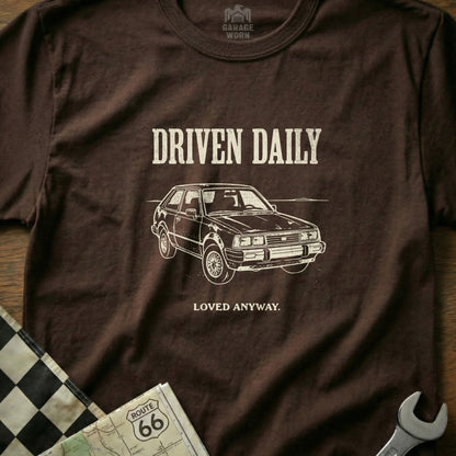 Brown t-shirt with "DRIVEN DAILY LOVED ANYWAY" text and a car graphic.