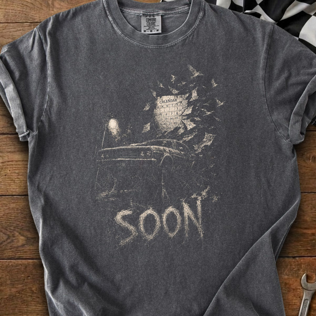 Dark t-shirt with a graphic of a car, calendar, and the word "SOON".