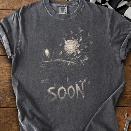 Dark t-shirt with a graphic of a car, calendar, and the word "SOON".