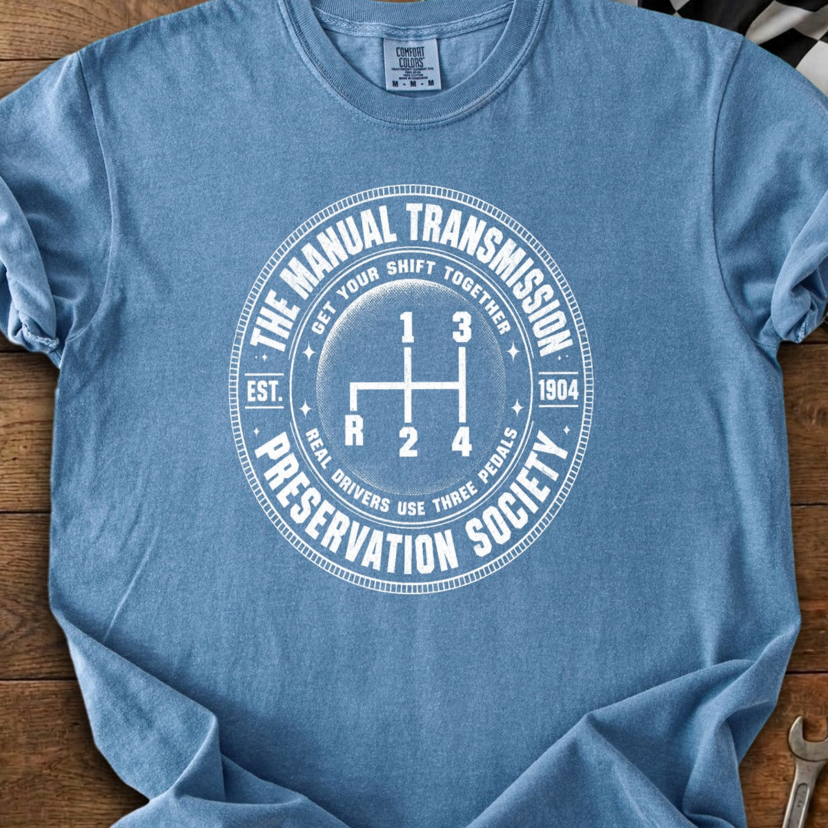 Blue t-shirt with white circular graphic showing a manual gear shift pattern and text.