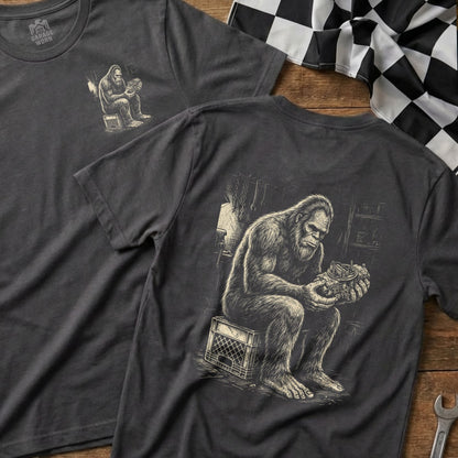 Bigfoot sitting on a crate, examining a carburetor on a dark t-shirt.