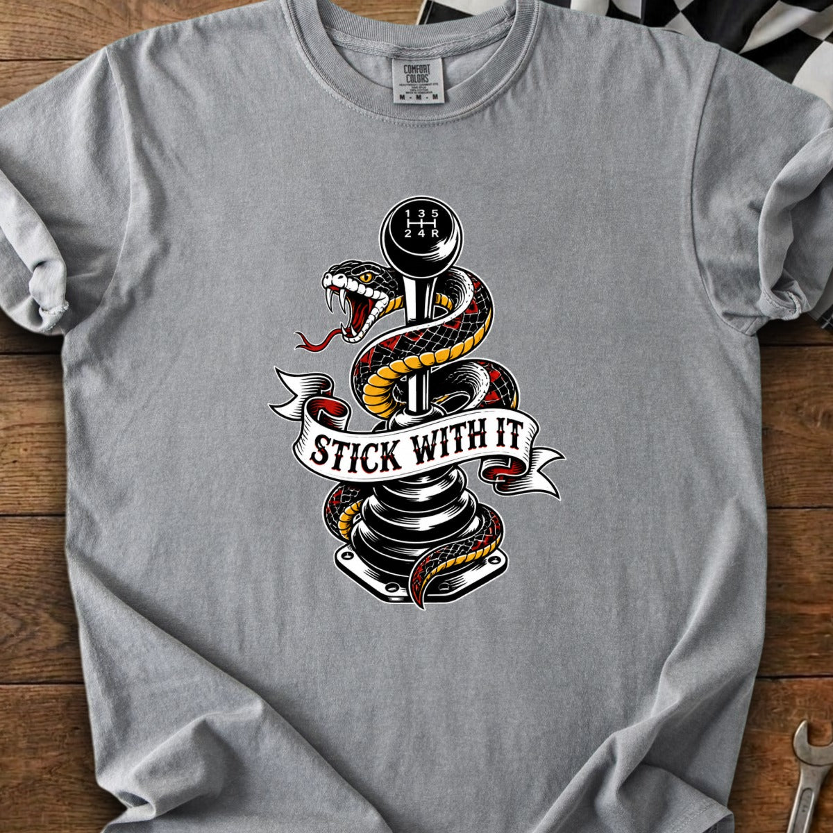 Snake wrapped around manual transmission shift knob with "STICK WITH IT" banner on grey t-shirt.