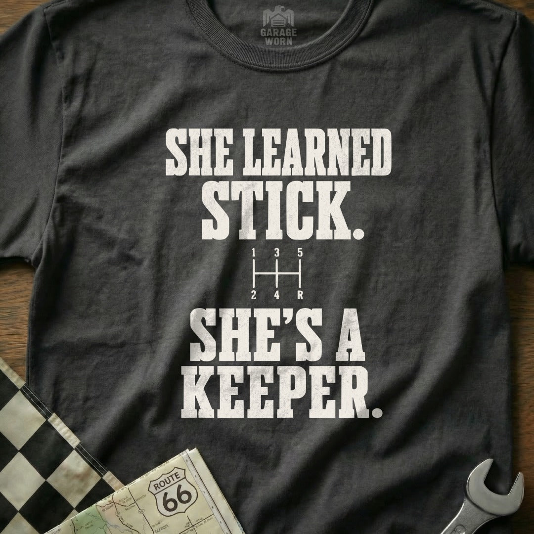 Dark t-shirt with white text "SHE LEARNED STICK. SHE'S A KEEPER."