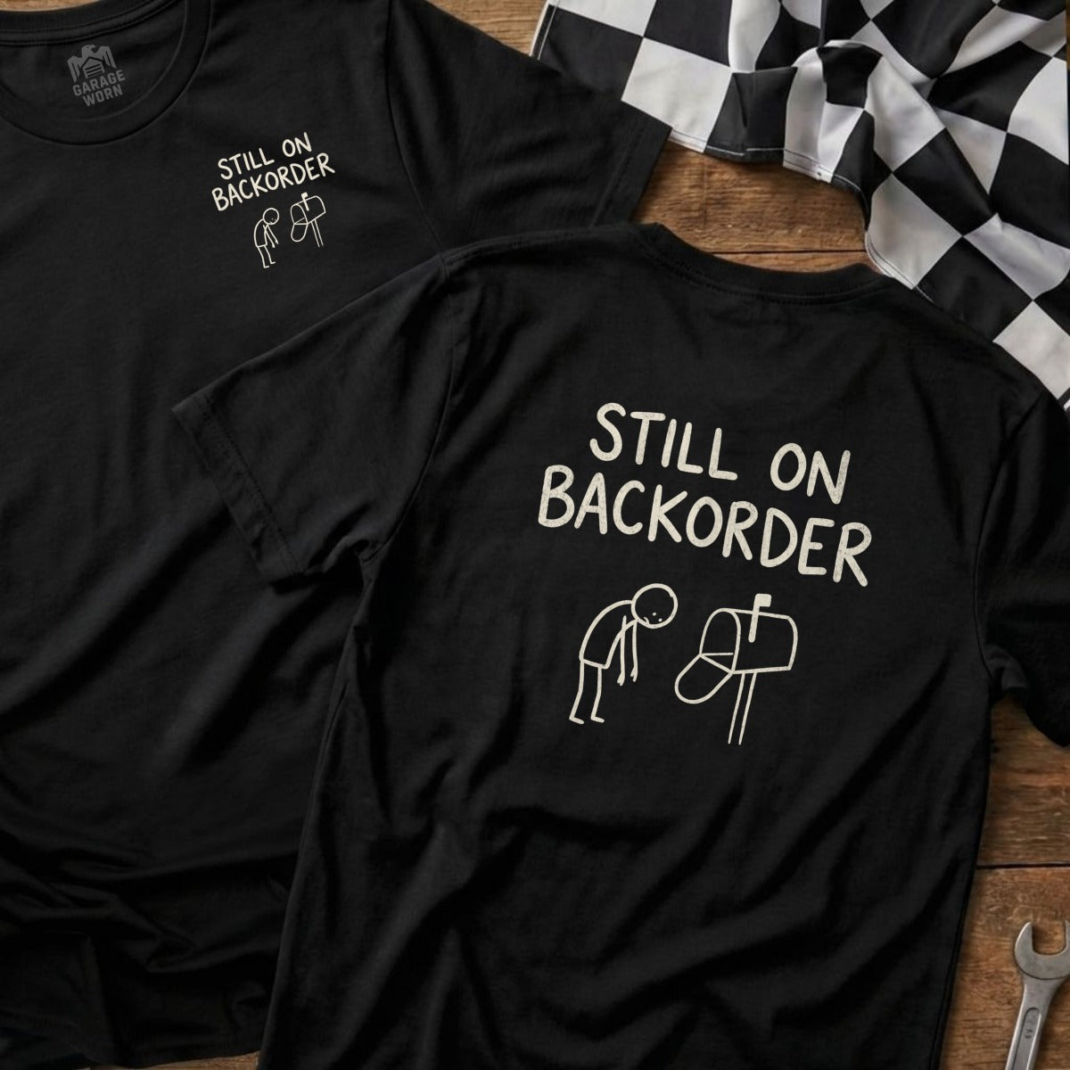 Black t-shirt with white text "Still On Backorder" and stick figure by a mailbox.