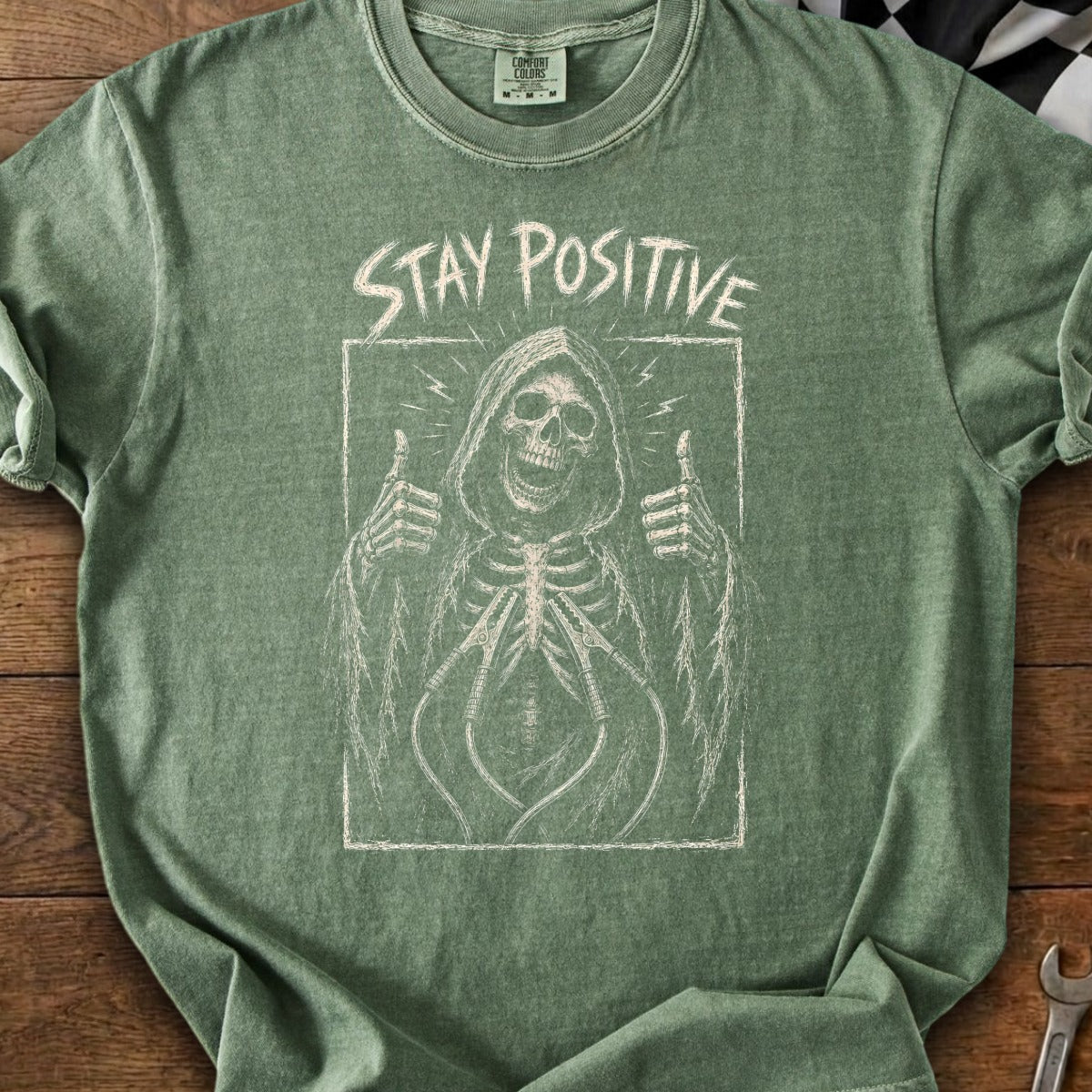 Green t-shirt with a chalk-style graphic of a skeleton giving a thumbs up with "STAY POSITIVE" text.