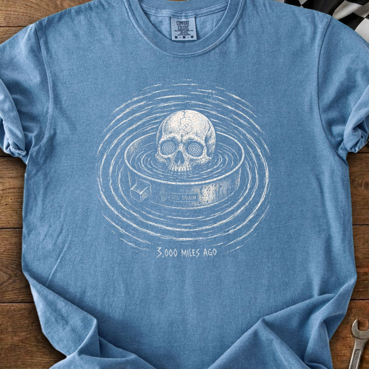 Skull in water with concentric rings, text "OIL DRAIN" and "3,000 MILES AGO".