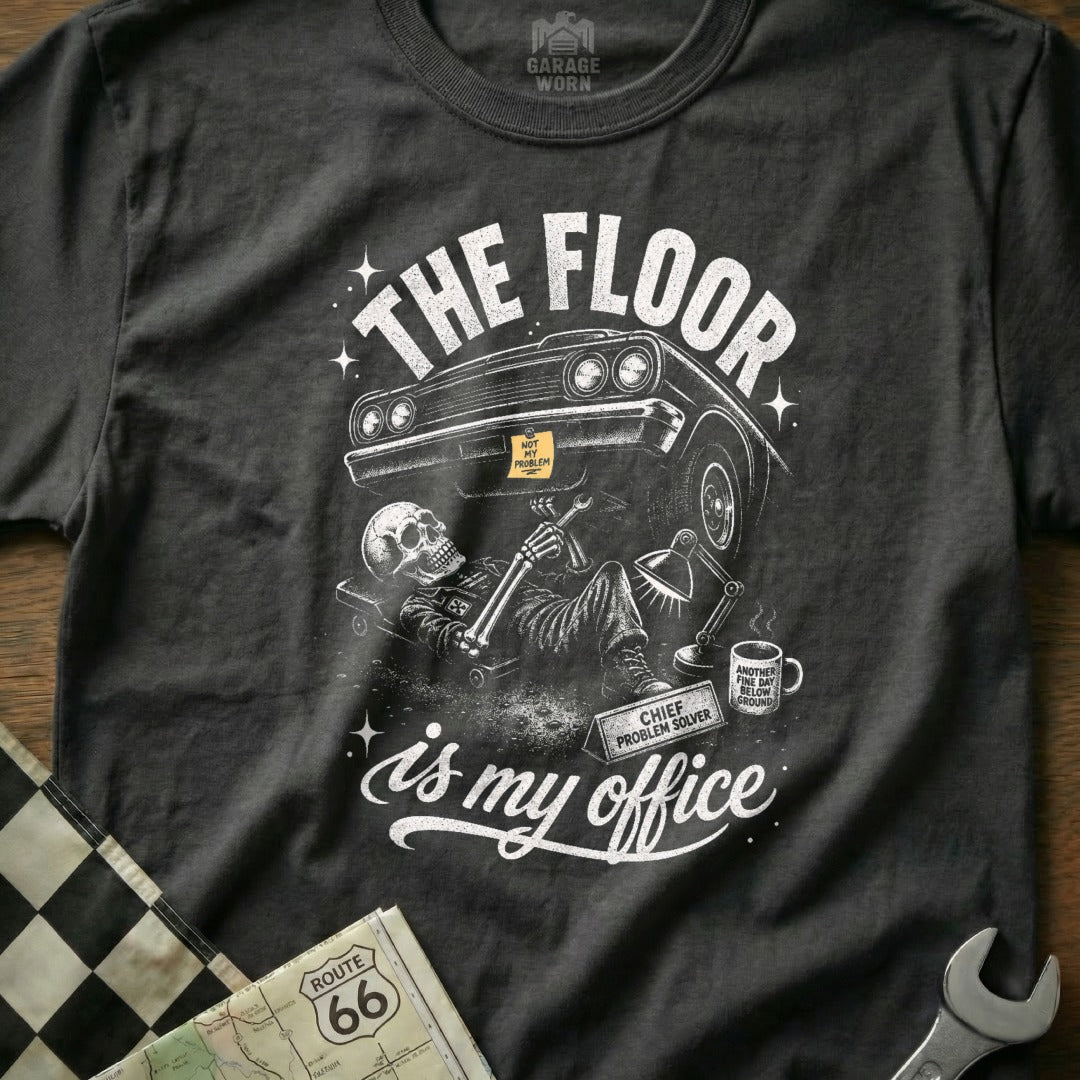 Dark gray t-shirt graphic of skeleton fixing car: "The Floor is My Office."