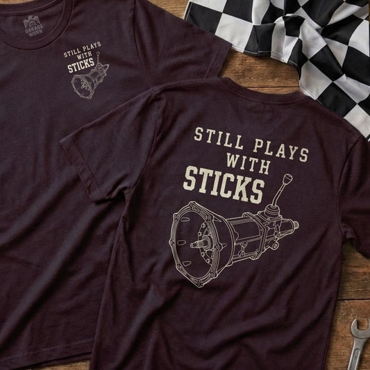 Maroon t-shirt with "Still Plays With Sticks" text and manual transmission graphic.