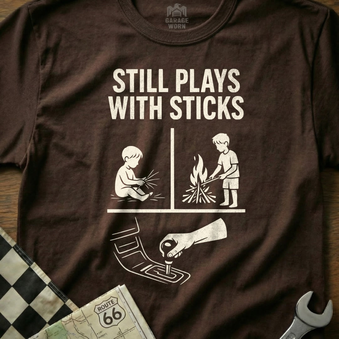 Brown t-shirt with white text and graphics: "Still Plays With Sticks" and contrasting stick figures.