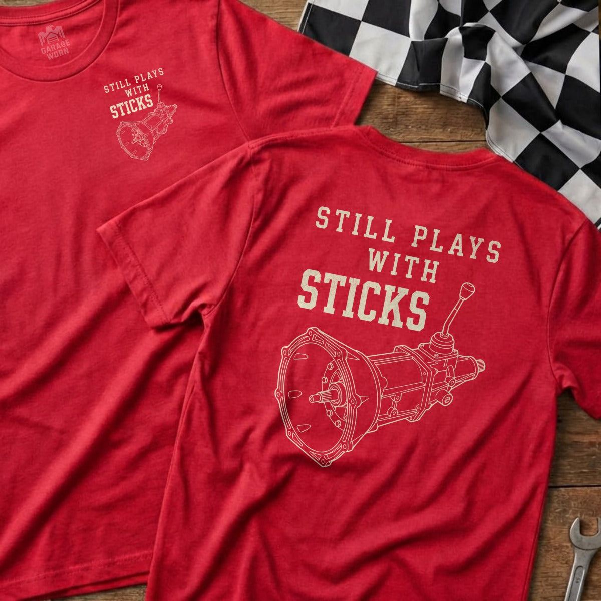 Two red t-shirts with "Still Plays With Sticks" and a drawing of a manual transmission.