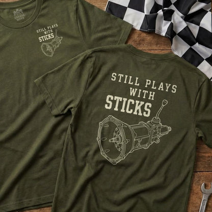 Two olive green t-shirts showing a manual transmission graphic and "STILL PLAYS WITH STICKS".