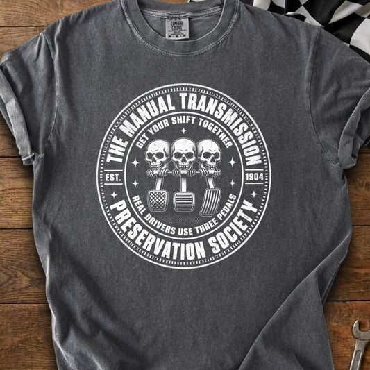 Graphic tee with skull graphic holding manual transmission pedals, "The Manual Transmission Preservation Society" text.