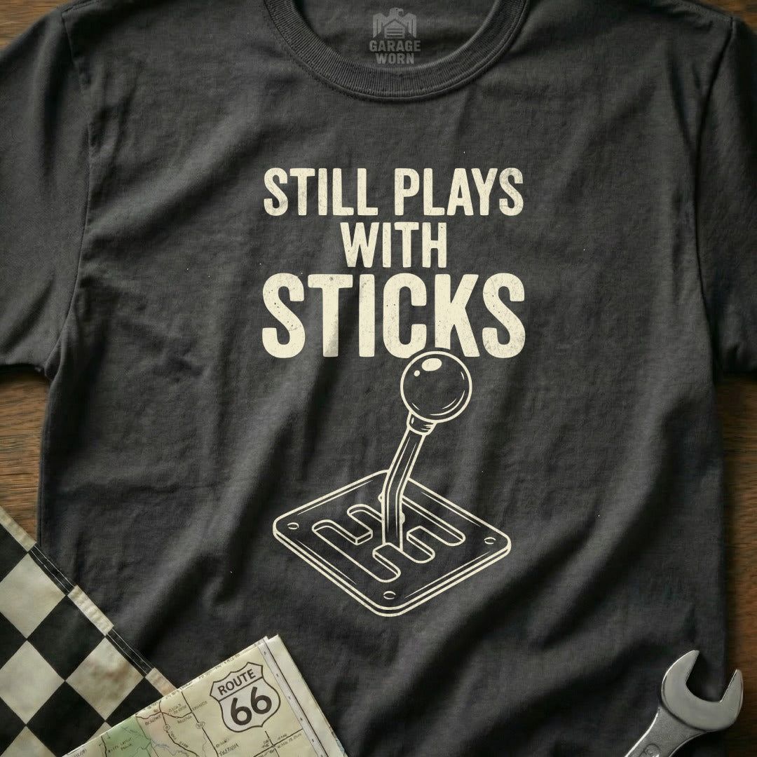 Dark t-shirt with "Still Plays With Sticks" and a manual shift knob graphic.