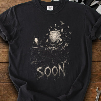 Black t-shirt with distressed graphic of a car, calendar, and circling birds, with "SOON" text.