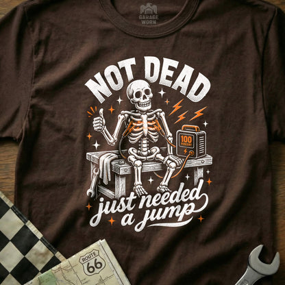 Brown t-shirt graphic with skeleton getting a jump start, "NOT DEAD just needed a jump".