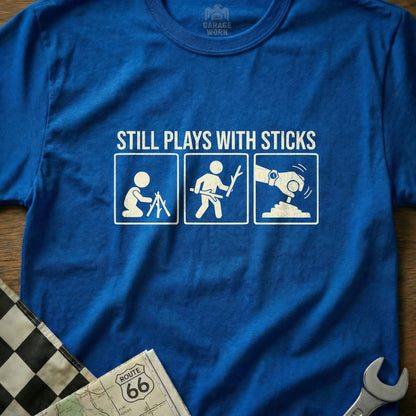 Blue t-shirt with "Still Plays With Sticks" text and three stick figure illustrations.