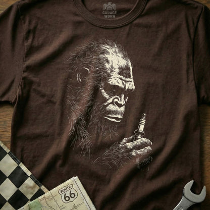 Brown t-shirt with a sketch of Bigfoot holding a spark plug, near a checkered flag.