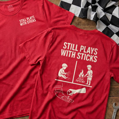 Red t-shirt with "STILL PLAYS WITH STICKS" text and graphic of child playing with sticks.