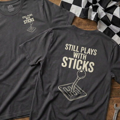 Dark grey t-shirt with "Still Plays with Sticks" and a manual transmission shifter graphic.