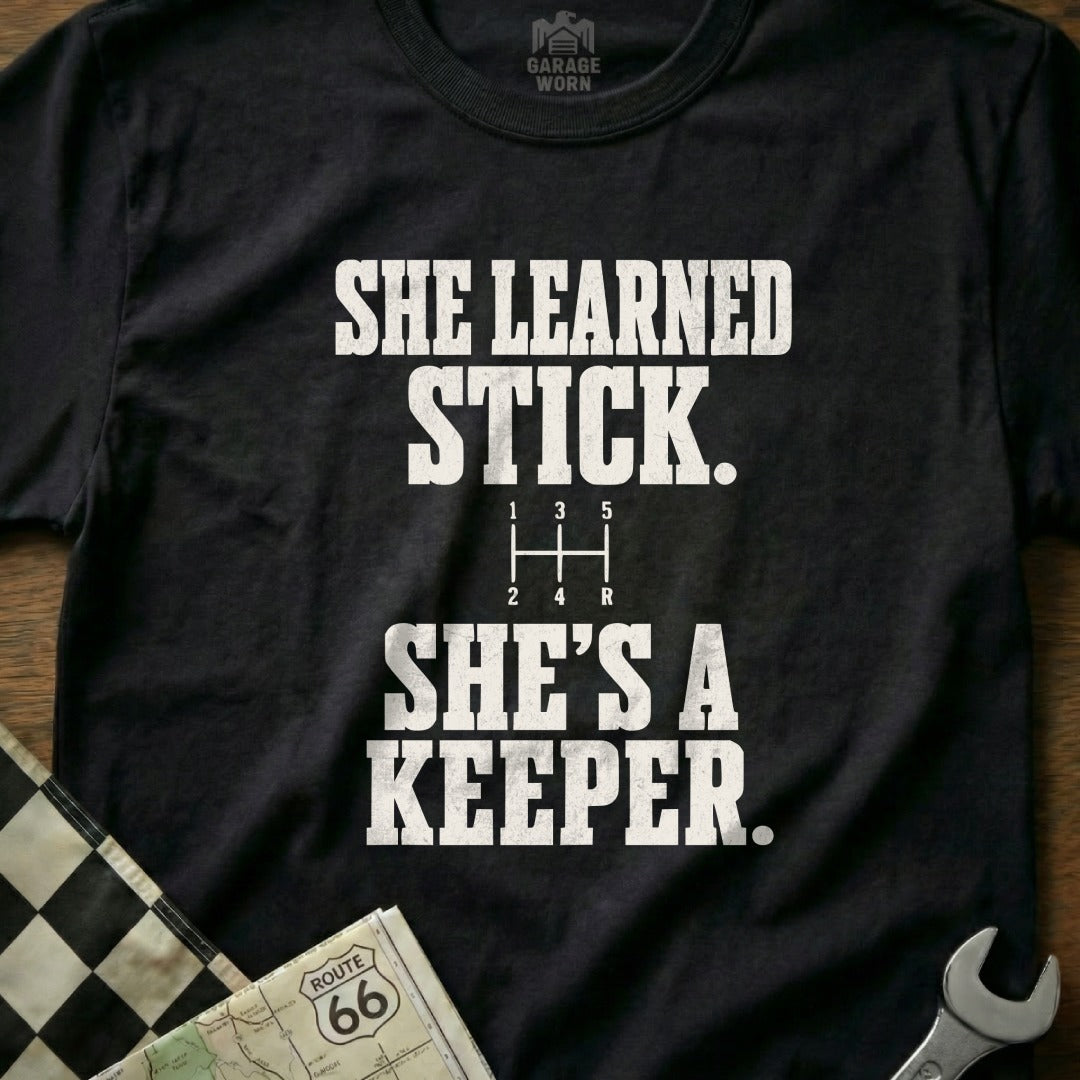 Black t-shirt with white text: "SHE LEARNED STICK. SHE'S A KEEPER."