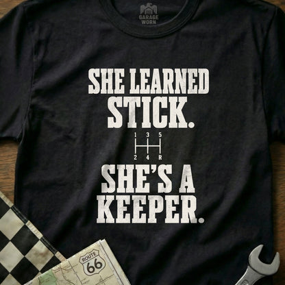 Black t-shirt with white text: "SHE LEARNED STICK. SHE'S A KEEPER."