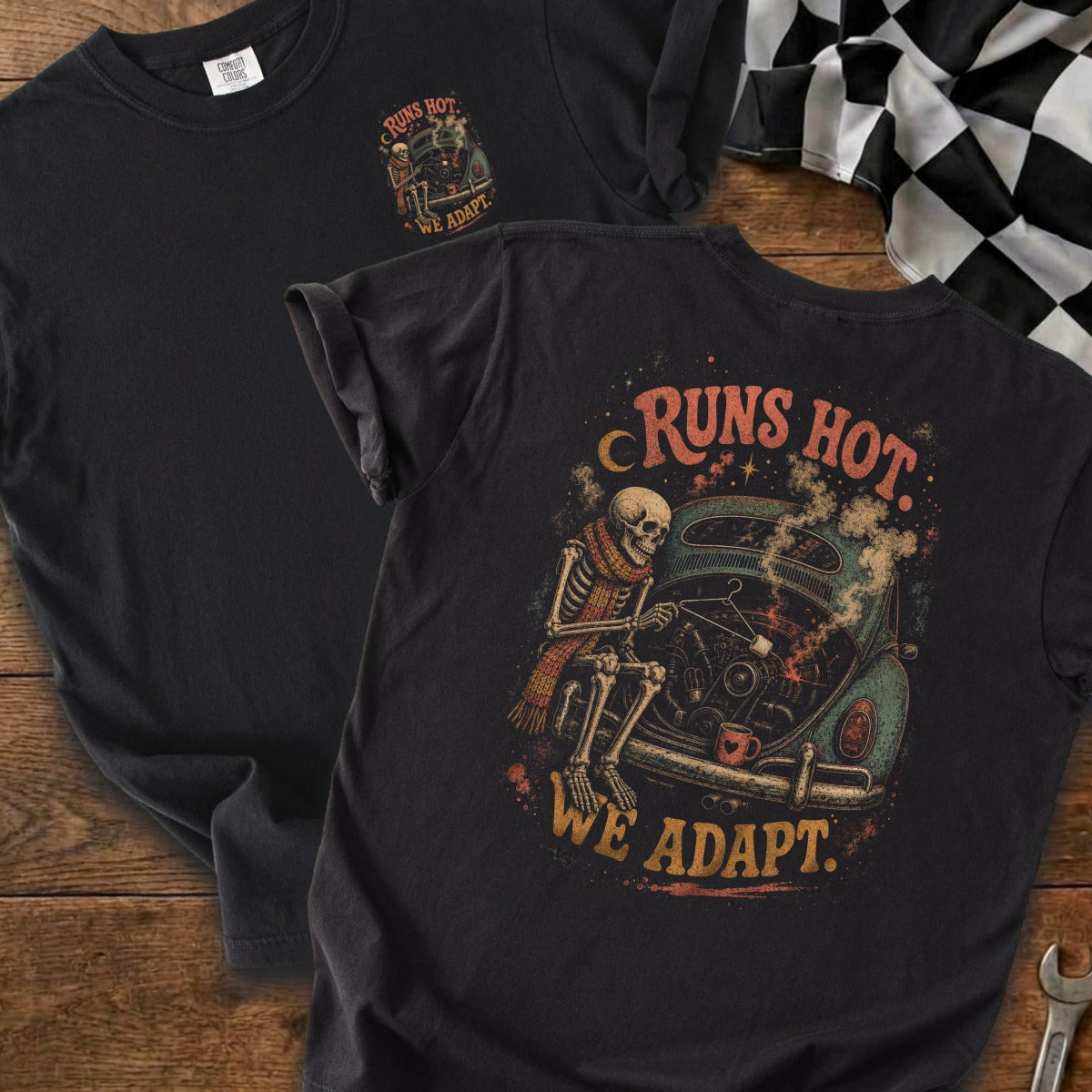 Dark t-shirt with skeleton working on car graphic; text reads "RUNS HOT. WE ADAPT."