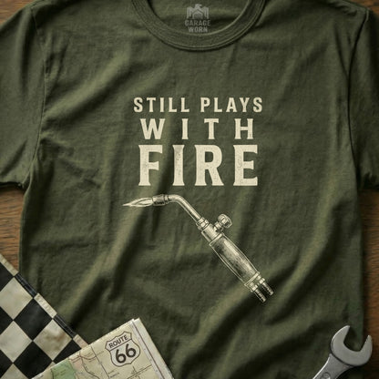 Green t-shirt with "Still Plays With Fire" text and a welding torch.