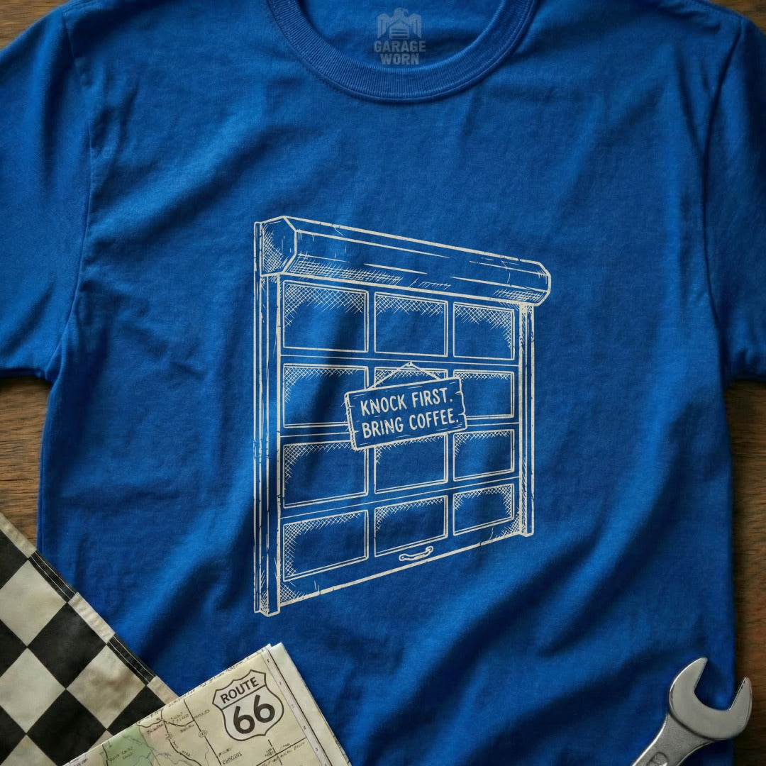 Blue t-shirt with a drawing of a garage door that reads "Knock First, Bring Coffee."