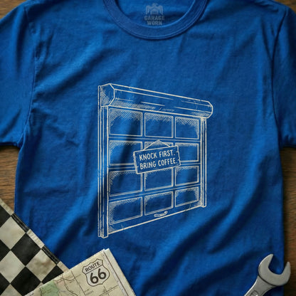 Blue t-shirt with a drawing of a garage door that reads "Knock First, Bring Coffee."