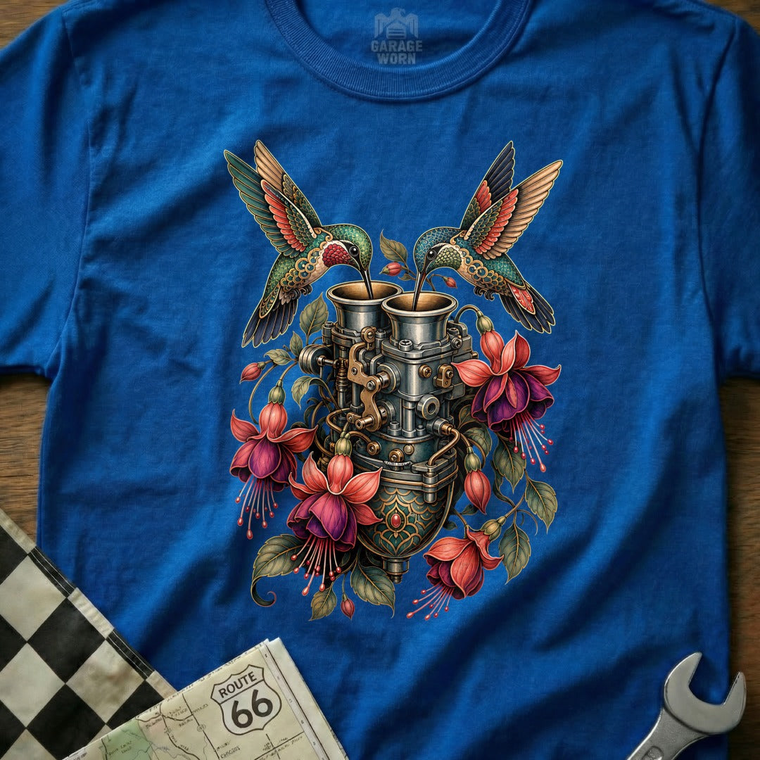 Two hummingbirds drink from a vintage carburetor surrounded by fuchsia flowers.