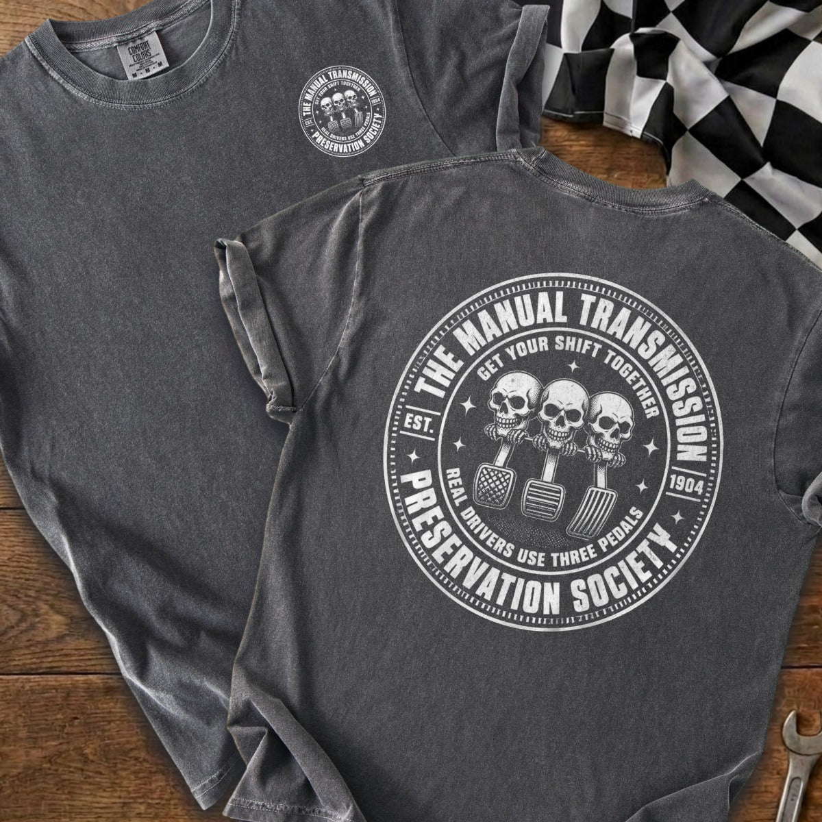 Dark gray t-shirt with circular graphic of three skulls on clutch, brake, and accelerator pedals reading "The Manual Transmission Preservation Society".