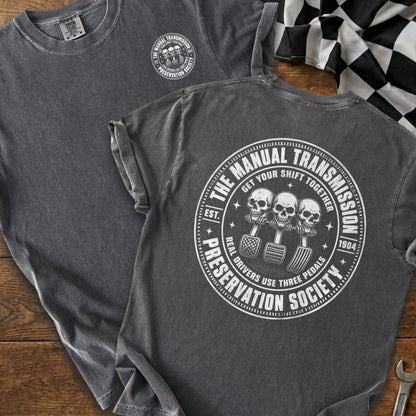 Dark gray t-shirt with circular graphic of three skulls on clutch, brake, and accelerator pedals reading "The Manual Transmission Preservation Society".