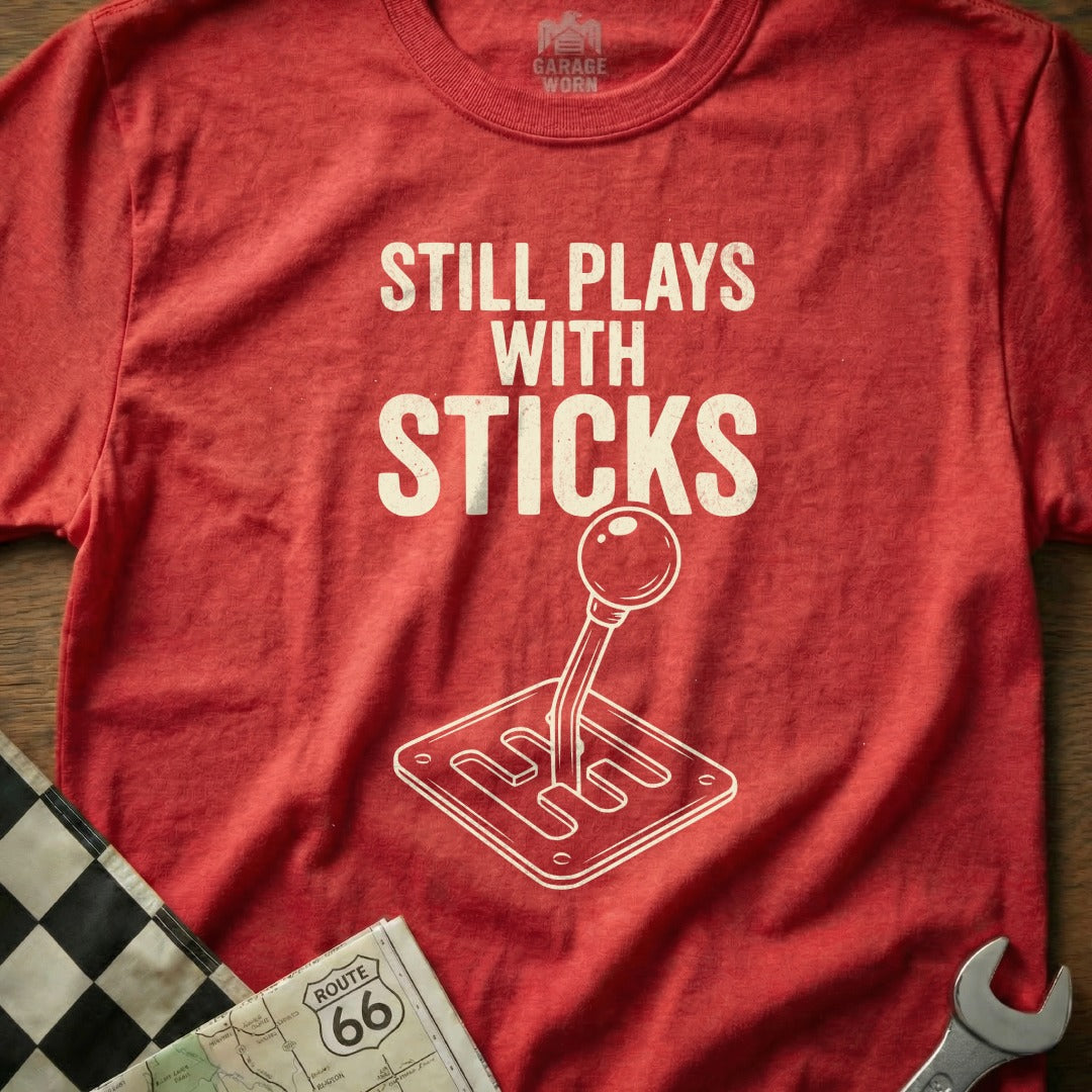 Red t-shirt with "Still Plays With Sticks" text and a gear shift graphic.